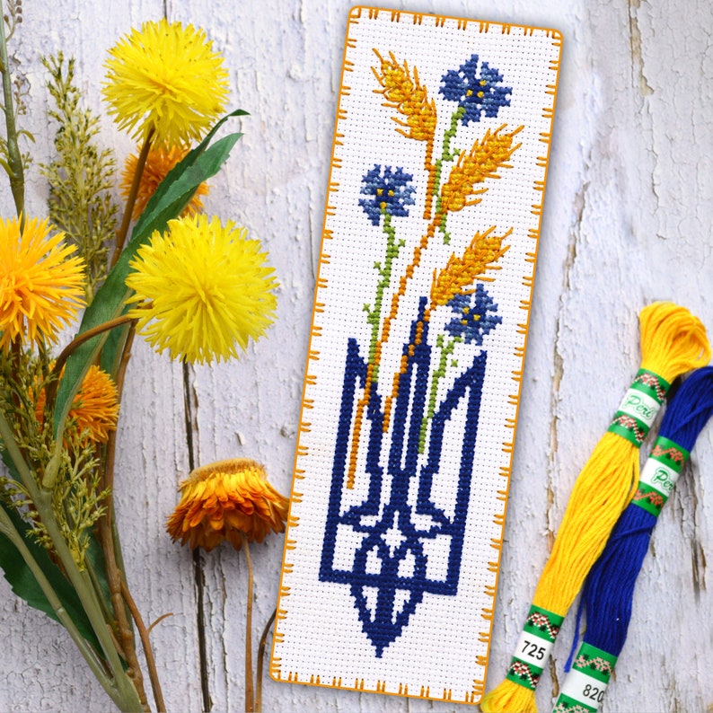 May include: Cross stitch bookmark featuring a blue trident, wheat stalks, and blue flowers on a white background. The bookmark is surrounded by a yellow border.