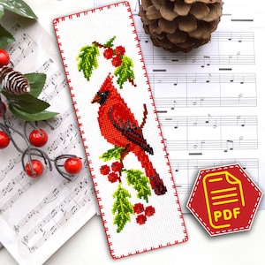 May include: A rectangular cross-stitch bookmark featuring a vibrant red cardinal bird perched on a branch with green leaves and red berries. The design is stitched on white fabric with a red stitched border. A red hexagonal PDF icon is visible.