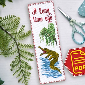 May include: A white cross-stitch bookmark with red stitching around the edges. The bookmark features a green alligator with a yellow belly swimming in blue water. A weeping willow tree is in the background. The text "A long time ago" is stitched in black thread at the top of the bookmark.