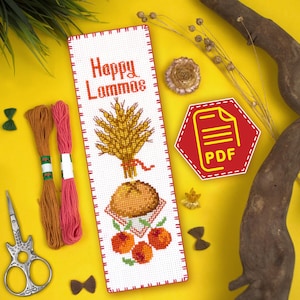 Puede incluir: Un marcapáginas de punto de cruz blanco con un borde rojo. El marcapáginas presenta una gavilla de trigo, una hogaza de pan y tres manzanas rojas. El texto "Happy Lammas" está bordado en rojo en la parte superior del marcapáginas.