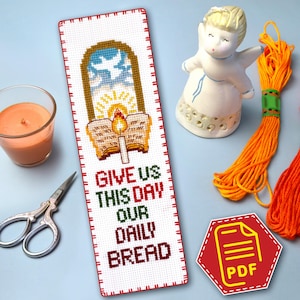 May include: A handmade cross-stitch bookmark with the text "GIVE US THIS DAY OUR DAILY BREAD". The bookmark features a window design with a dove, sun, and open book with a candle. A small angel figurine and orange thread are also visible.