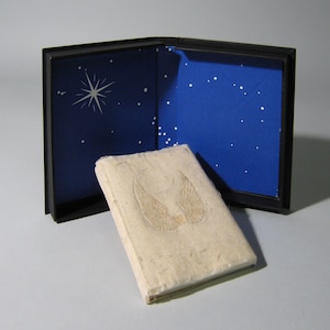 May include: A black box with a blue interior decorated with white stars. The box is open and a cream-colored journal with a raised image of wings is inside.