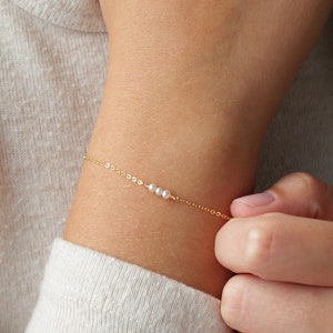 May include: A delicate gold chain bracelet with three small white pearls. The bracelet is worn on a person's wrist.