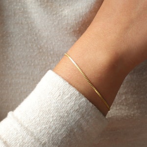 May include: A delicate gold bracelet with a square link chain is worn on a wrist. The bracelet is a simple, elegant piece of jewelry. The person is wearing a light beige sweater.