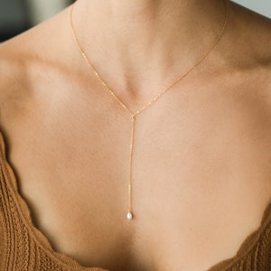 Peut inclure: Un collier délicat en chaîne dorée avec un pendentif en perle unique. Le collier est porté par une personne avec un haut marron.