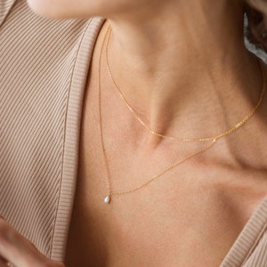 May include: A gold chain necklace with a small pearl pendant. The necklace is layered with a second, thinner gold chain.