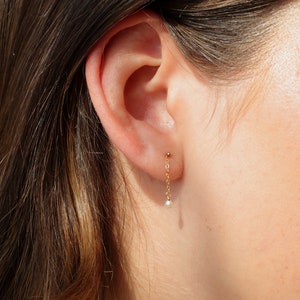 May include: Gold chain earrings with a small pearl dangling at the end. The earrings are worn in a person's ear.