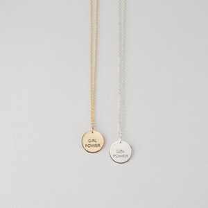 Puede incluir: Dos collares con un colgante redondo que dice "Girl Power" en may&uacute;sculas. Un collar tiene una cadena y un colgante dorados, y el otro tiene una cadena y un colgante plateados.