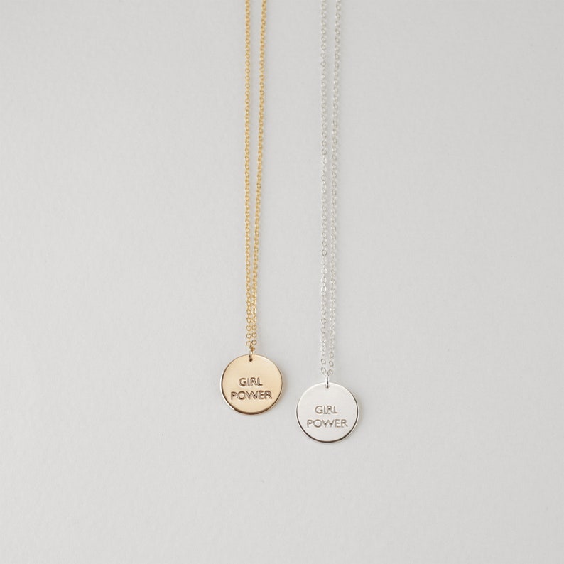 Puede incluir: Dos collares con un colgante redondo que dice "Girl Power" en may&uacute;sculas. Un collar tiene una cadena y un colgante dorados, y el otro tiene una cadena y un colgante plateados.
