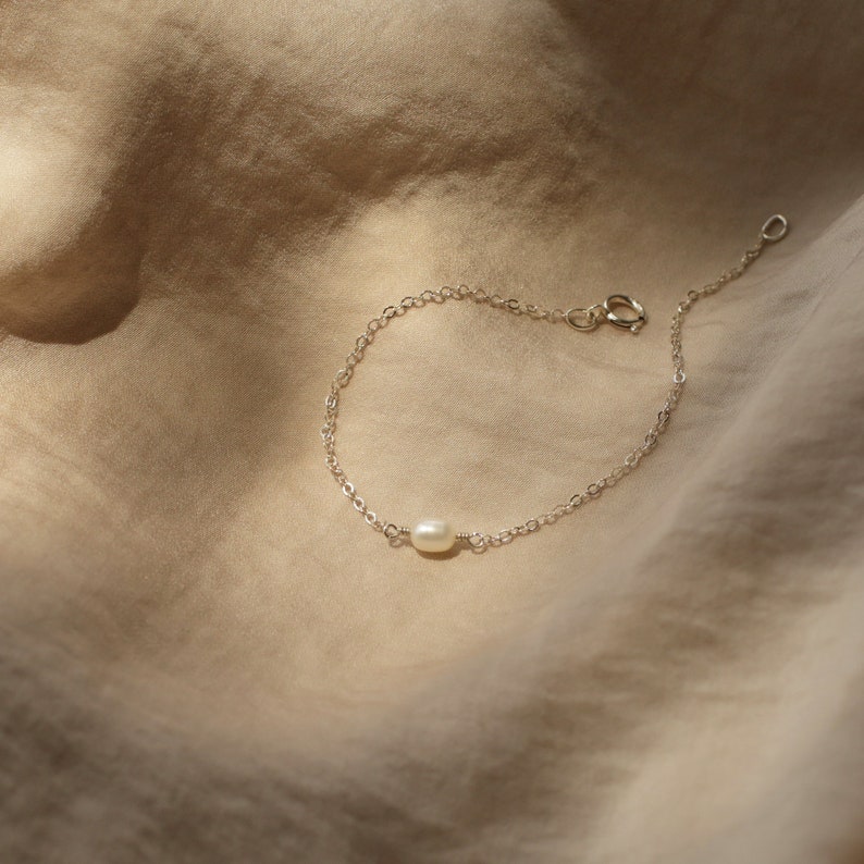 May include: A delicate silver chain bracelet with a single white pearl charm. The bracelet is on a light brown fabric background.