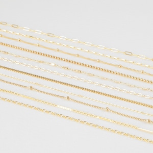 May include: A collection of gold chains of varying lengths and styles, including cable, curb, and box chains. The chains are arranged in a staggered pattern on a white background.