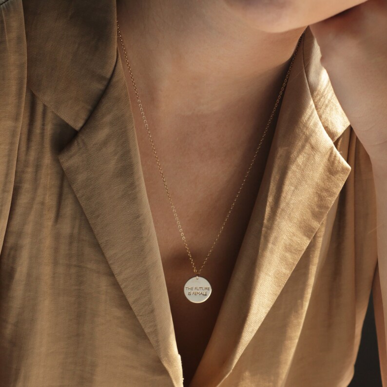 Puede incluir: Un collar de cadena dorada con un colgante redondo que dice "THE FUTURE IS FEMALE".