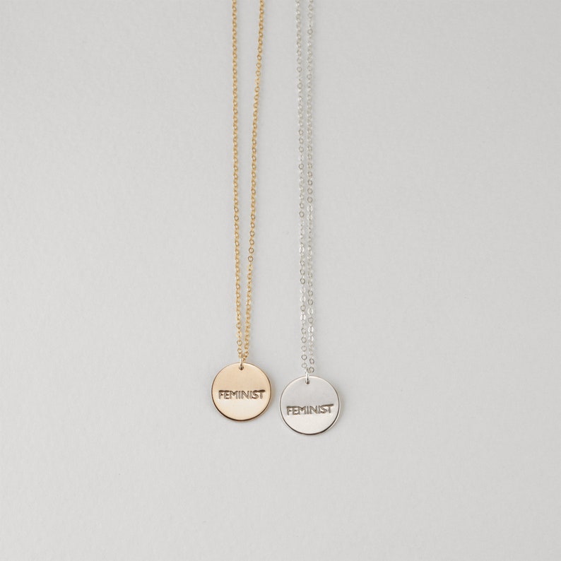 Puede incluir: Dos delicados collares con colgantes circulares que dicen "FEMINIST" en una fuente sencilla. Un collar tiene una cadena y un colgante dorados, mientras que el otro tiene una cadena y un colgante plateados.