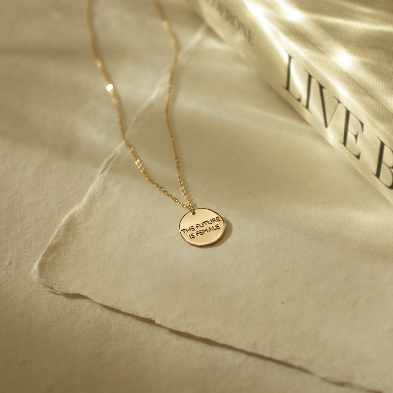 Puede incluir: Un collar de oro con un colgante redondo que dice "THE FUTURE IS FEMALE".