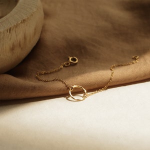 May include: A delicate gold chain bracelet with a simple circle pendant. The bracelet is lying on a brown fabric background.