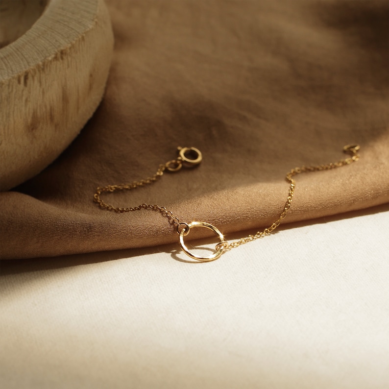May include: A delicate gold chain bracelet with a simple circle pendant. The bracelet is lying on a brown fabric background.