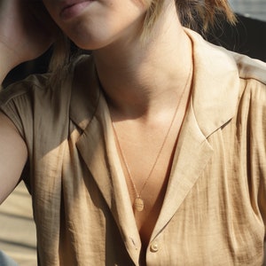 Puede incluir: Un collar de cadena dorada con un colgante redondo que dice "Sigue adelante, t&uacute; puedes". El collar lo lleva una persona con el pelo corto y rubio que lleva una camisa marr&oacute;n claro.