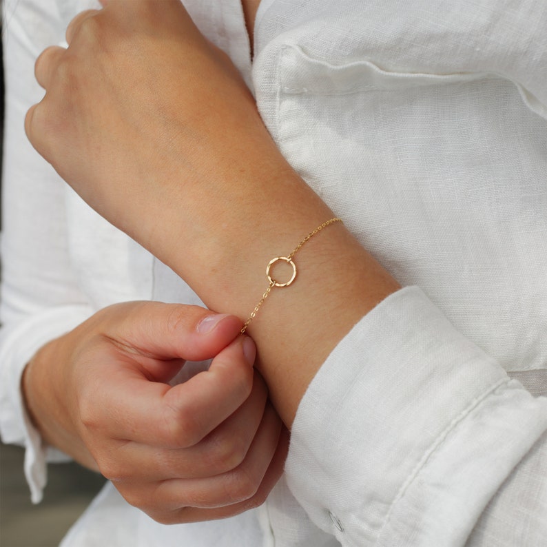 May include: A delicate gold chain bracelet with a simple circle pendant. The bracelet is shown on a person's wrist.
