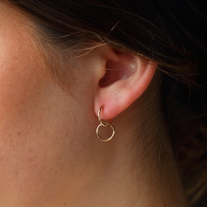 May include: Gold hoop earrings with a smaller hoop hanging from the larger hoop. The earrings are worn in a person's ear.