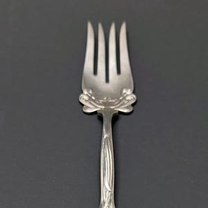 May include: A silver-toned metal fork featuring four tines and an ornate handle. The handle displays intricate embossed floral and leaf motifs, with a central shield-shaped cartouche containing an engraved letter E at the base of the stem.