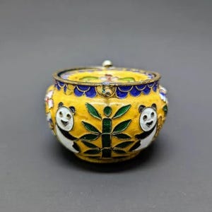 Pu&ograve; includere: Una piccola scatola rotonda in cloisonn&eacute; gialla con un coperchio. La scatola &egrave; decorata con orsi panda bianchi e neri, bamb&ugrave; verde e fiori blu e bianchi.