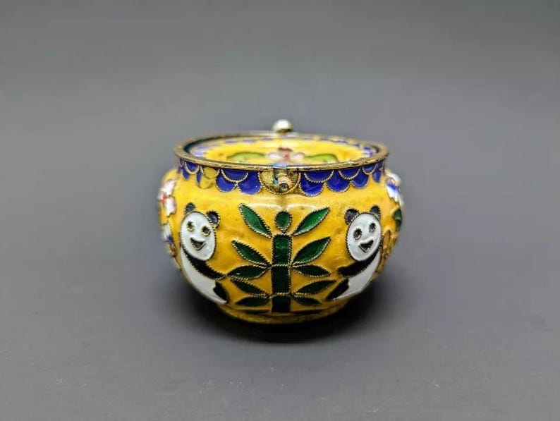 Pu&ograve; includere: Una piccola scatola rotonda in cloisonn&eacute; gialla con un coperchio. La scatola &egrave; decorata con orsi panda bianchi e neri, bamb&ugrave; verde e fiori blu e bianchi.
