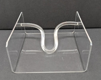 May include: Clear acrylic napkin holder with a curved handle design. The holder has a rectangular base and two upright sides. The clear material allows for visibility of the napkins. The holder is designed to keep napkins organized and accessible.