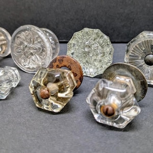May include: A collection of clear glass door knobs with various designs, including faceted, floral, and round shapes. Some knobs have metal hardware. The knobs are arranged on a dark gray surface.