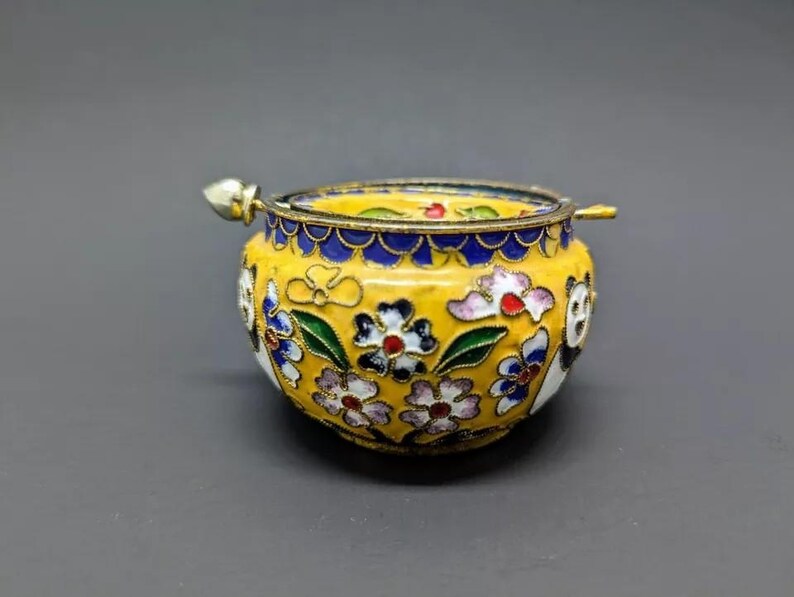 Pu&ograve; includere: Una piccola scatola portaoggetti in cloisonn&eacute; gialla con un motivo floreale e un coperchio con un piccolo pomo. La scatola &egrave; decorata con fiori bianchi, blu, verdi e rossi.