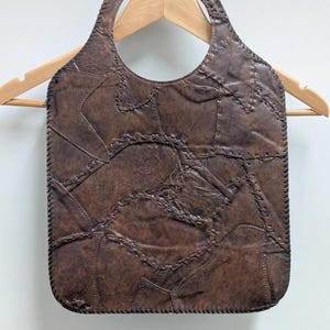 May include: Brown leather handbag with a unique patchwork design. The bag features a rounded handle and is constructed from various leather pieces stitched together. The bag is hanging on a wooden hanger against a white background.