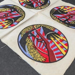 May include: Four square patches with a circular design featuring a rifle, wheat, and lightning bolts in red, blue, and yellow. The patches are on a cream-colored fabric, with a black background.