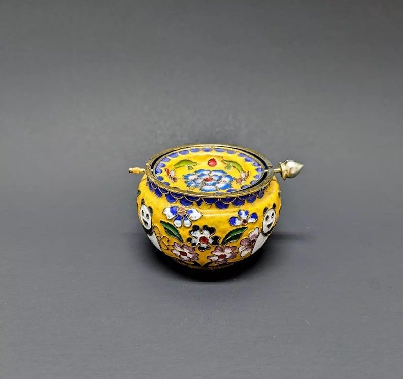 Pu&ograve; includere: Una piccola scatola rotonda in smalto cloisonn&eacute; gialla con un motivo floreale e un coperchio in argento. Il coperchio ha un piccolo pomo in argento sulla parte superiore.
