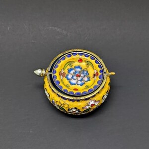Pu&ograve; includere: Una piccola scatola rotonda in smalto giallo con un motivo floreale. Il coperchio &egrave; aperto e presenta un motivo floreale blu e bianco. La scatola ha una cerniera argentata e un pomello argentato.