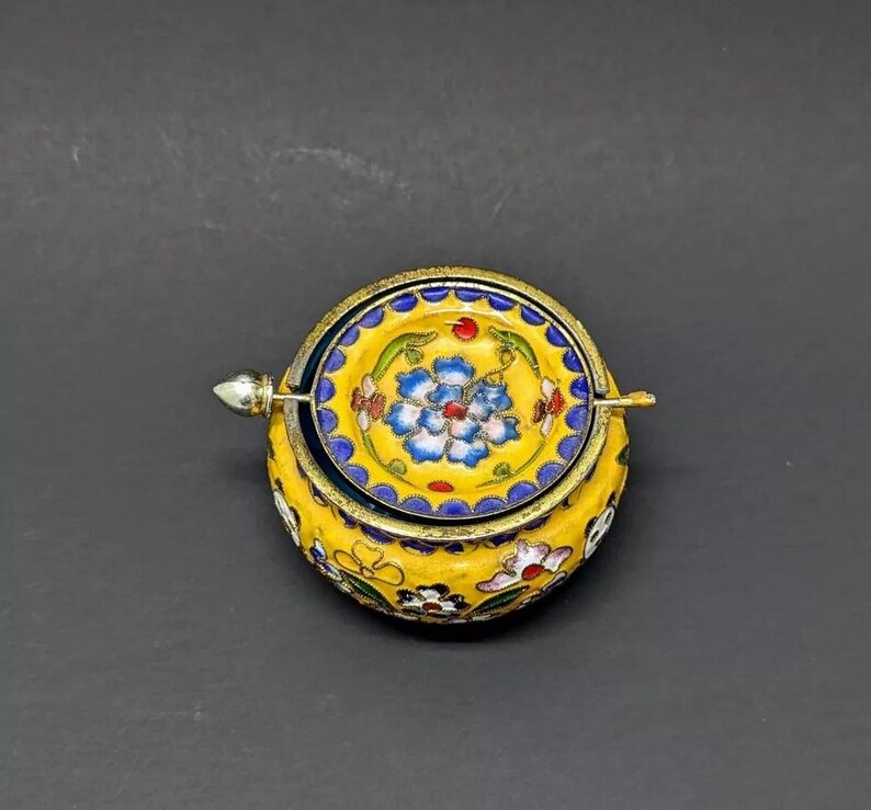 Pu&ograve; includere: Una piccola scatola rotonda in smalto giallo con un motivo floreale. Il coperchio &egrave; aperto e presenta un motivo floreale blu e bianco. La scatola ha una cerniera argentata e un pomello argentato.