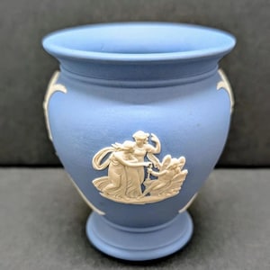 May include: A small, light blue ceramic vase with a round base and a wide opening. The vase features white relief figures of classical women. The vase is approximately 4 inches tall and is a decorative item.