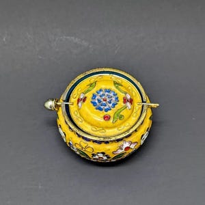 Pu&ograve; includere: Una piccola scatola rotonda gialla con un coperchio a cerniera. La scatola &egrave; decorata con motivi floreali colorati in blu, rosso, verde e bianco. Il coperchio ha una piccola maniglia argentata.