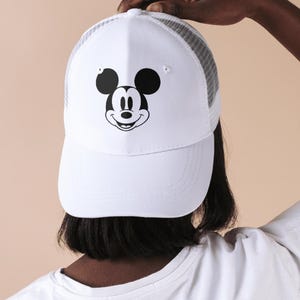 May include: White baseball cap featuring a black and white Mickey Mouse graphic on the front. The cap has a white front panel and a gray mesh back. The brim is curved, and the cap is being held up.