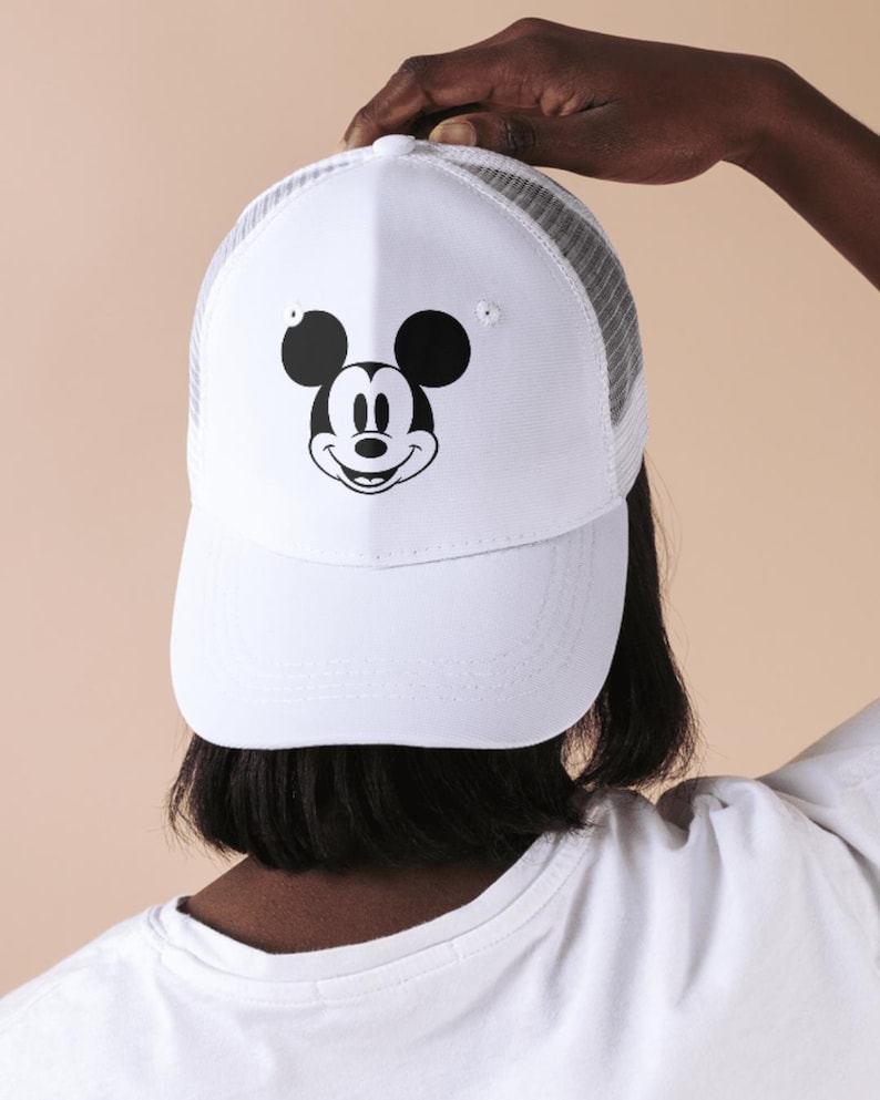 May include: White baseball cap featuring a black and white Mickey Mouse graphic on the front. The cap has a white front panel and a gray mesh back. The brim is curved, and the cap is being held up.
