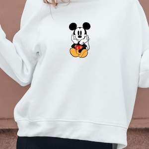 May include: White hooded sweatshirt featuring a graphic of Mickey Mouse. The cartoon character is depicted in a seated position with his hands under his chin. Mickey Mouse is wearing red shorts, yellow shoes, and white gloves.
