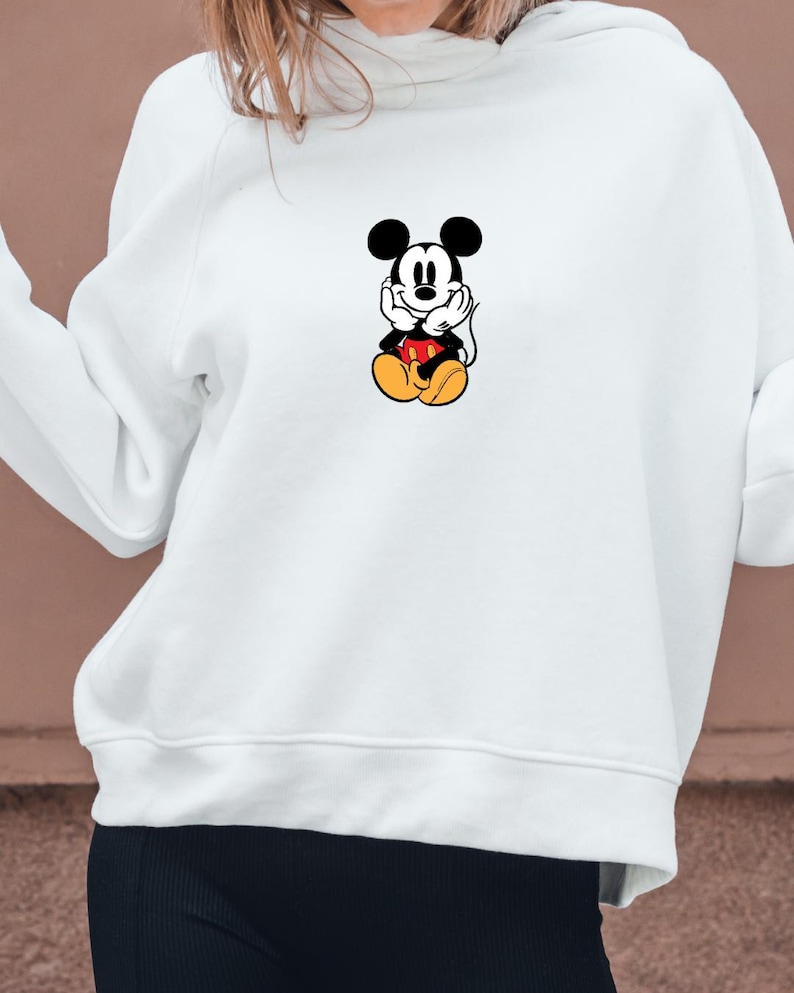 May include: White hooded sweatshirt featuring a graphic of Mickey Mouse. The cartoon character is depicted in a seated position with his hands under his chin. Mickey Mouse is wearing red shorts, yellow shoes, and white gloves.
