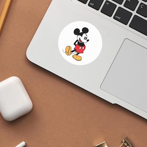 May include: A white circular sticker featuring a classic cartoon character, Mickey Mouse. Mickey is depicted in his iconic outfit: red shorts, yellow shoes, and black gloves. The sticker is placed on a laptop, with a brown surface in the background.