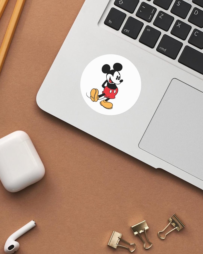 May include: A white circular sticker featuring a classic cartoon character, Mickey Mouse. Mickey is depicted in his iconic outfit: red shorts, yellow shoes, and black gloves. The sticker is placed on a laptop, with a brown surface in the background.