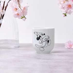 May include: White ceramic mug featuring a black and white illustration of Mickey Mouse in a wizard hat. The mug has a rounded shape and is set against a soft, neutral background with pink flowers.