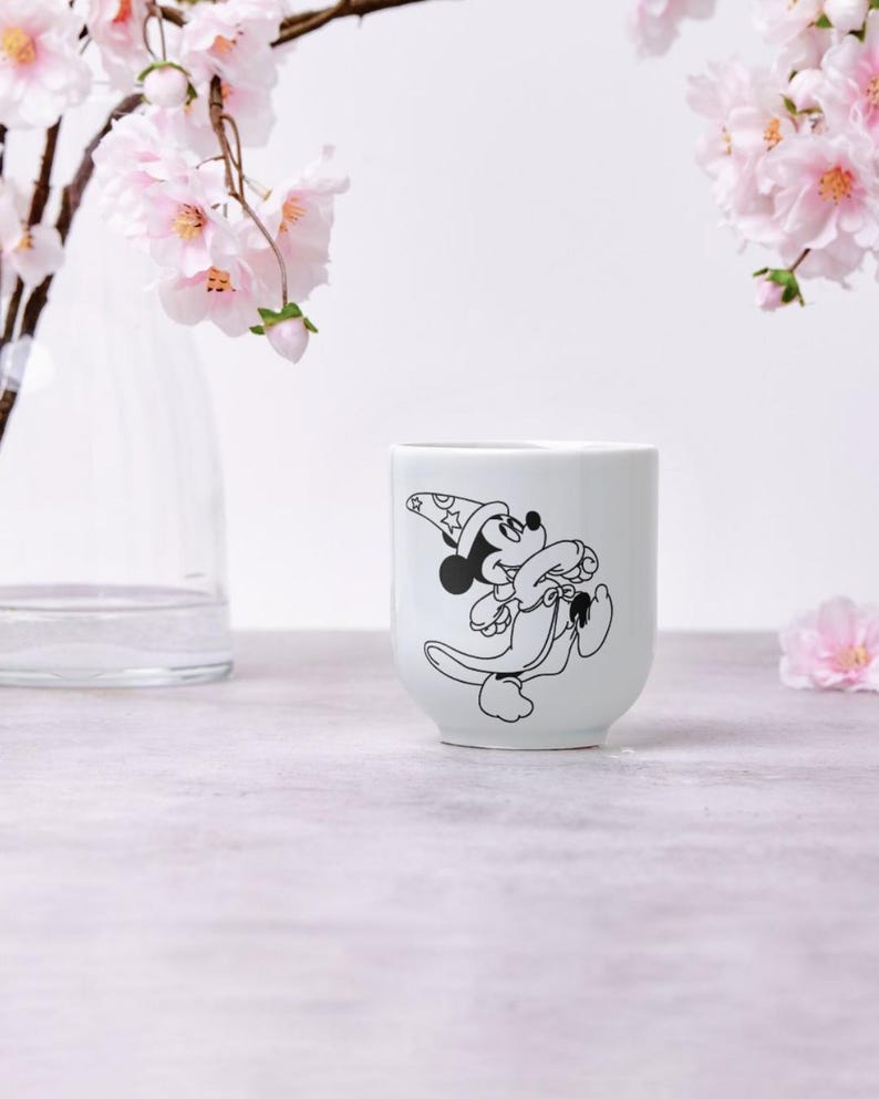 May include: White ceramic mug featuring a black and white illustration of Mickey Mouse in a wizard hat. The mug has a rounded shape and is set against a soft, neutral background with pink flowers.