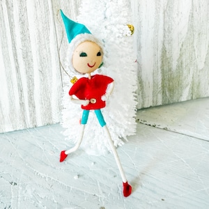 May include: A vintage Christmas elf ornament. The elf has a teal hat with white trim, a red shirt, turquoise legs, and red shoes. The elf is standing in front of a white, fluffy tree.