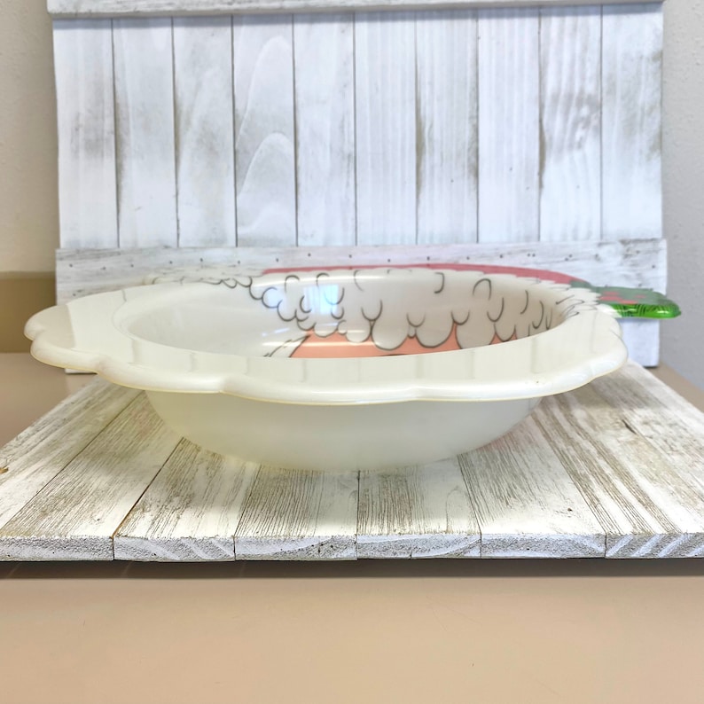 May include: A white plastic bowl with a pink and white strawberry design. The bowl has a scalloped edge and is perfect for serving snacks or fruit.