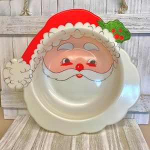 May include: A white plastic Santa Claus serving bowl with a red hat and green holly. The bowl is perfect for serving snacks or desserts during the holiday season.