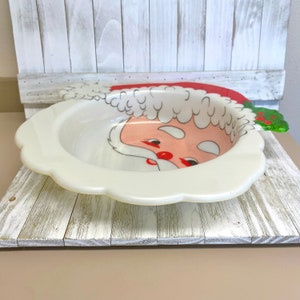May include: A white ceramic bowl with a Santa Claus face design. The bowl has a red hat, white beard, and black eyes. The bowl is on a white wooden surface.