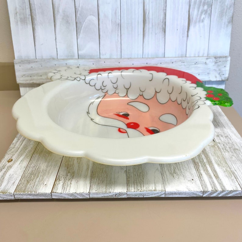 May include: A white ceramic bowl with a Santa Claus face design. The bowl has a red hat, white beard, and black eyes. The bowl is on a white wooden surface.