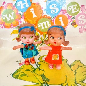 Könnte beinhalten: Zwei Vintage-Plastikpuppen mit braunen Zöpfen und blauen Stirnbändern. Eine Puppe trägt ein blaues Kleid, die andere ein orangefarbenes Kleid. Beide Puppen haben rote Schuhe und ausgestreckte Arme. Der Hintergrund zeigt bunte Blumen- und Buchstabenmuster.